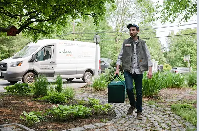 A driver carries a tote up a customer's garden path. A Walden Local branded delivery van waits at the curb.