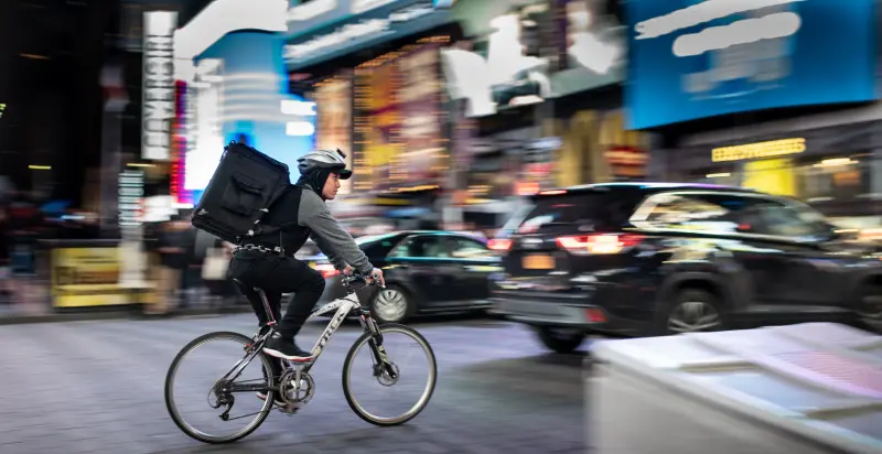 A cyclist wearing a square food delivery backpack on a busy city road at night. There is motion blur on the surrounding buildings, cars, and lights.