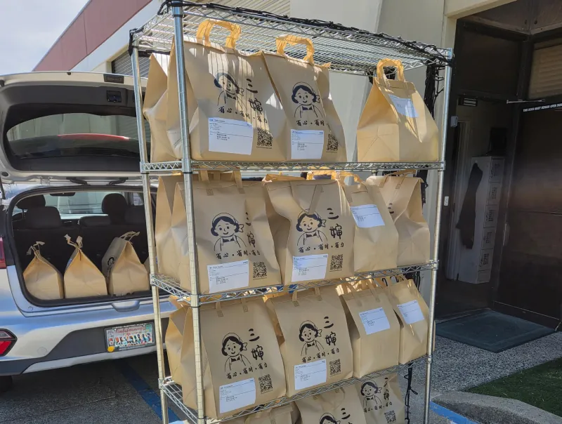 Branded Three Aunties totes are stacked on a wire rack, ready for loading and delivery to customers
