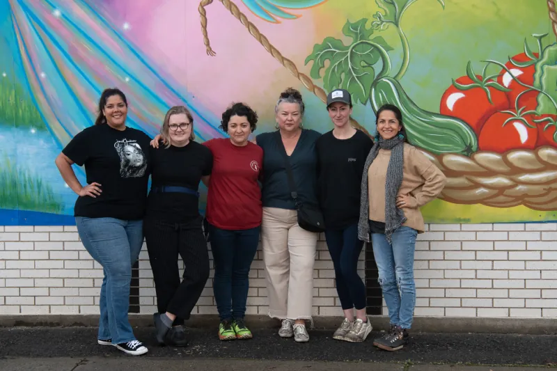Members of the Red Apron leadership pose with the artists of a colorful mural, which is partly visible in the background, showing images of freshly harvested cucumber and tomato in a basket.
