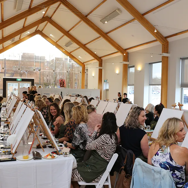 Group of mostly women seated at long tables in a bright room with wooden beams, each with blank canvases and paint supplies for a painting class or event.