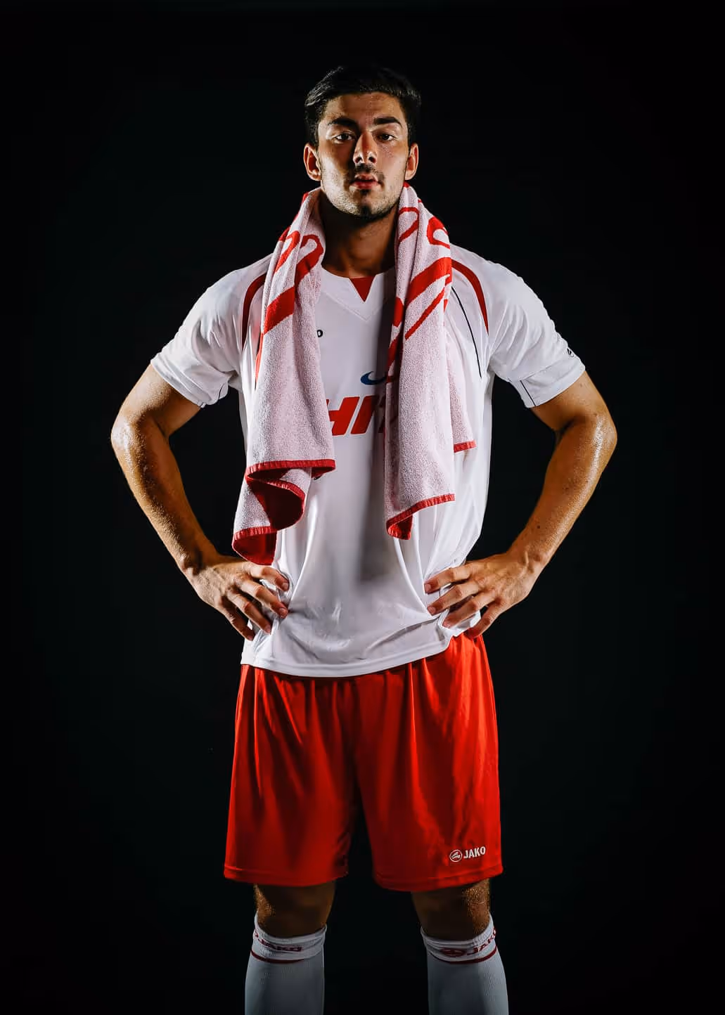 Mann in weißem Sporttrikot und roten Shorts mit Handtuch über den Schultern vor schwarzem Hintergrund.