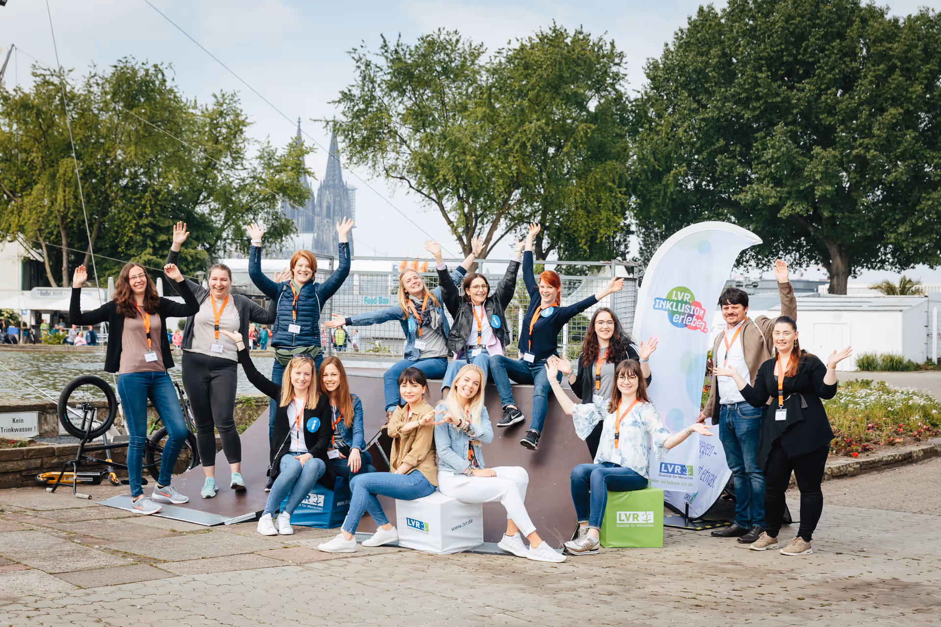 Gruppe von 15 Personen am Ufer mit erhobenen Händen, sitzend und stehend vor Bäumen und einem Fluss mit Kölner Dom im Hintergrund.