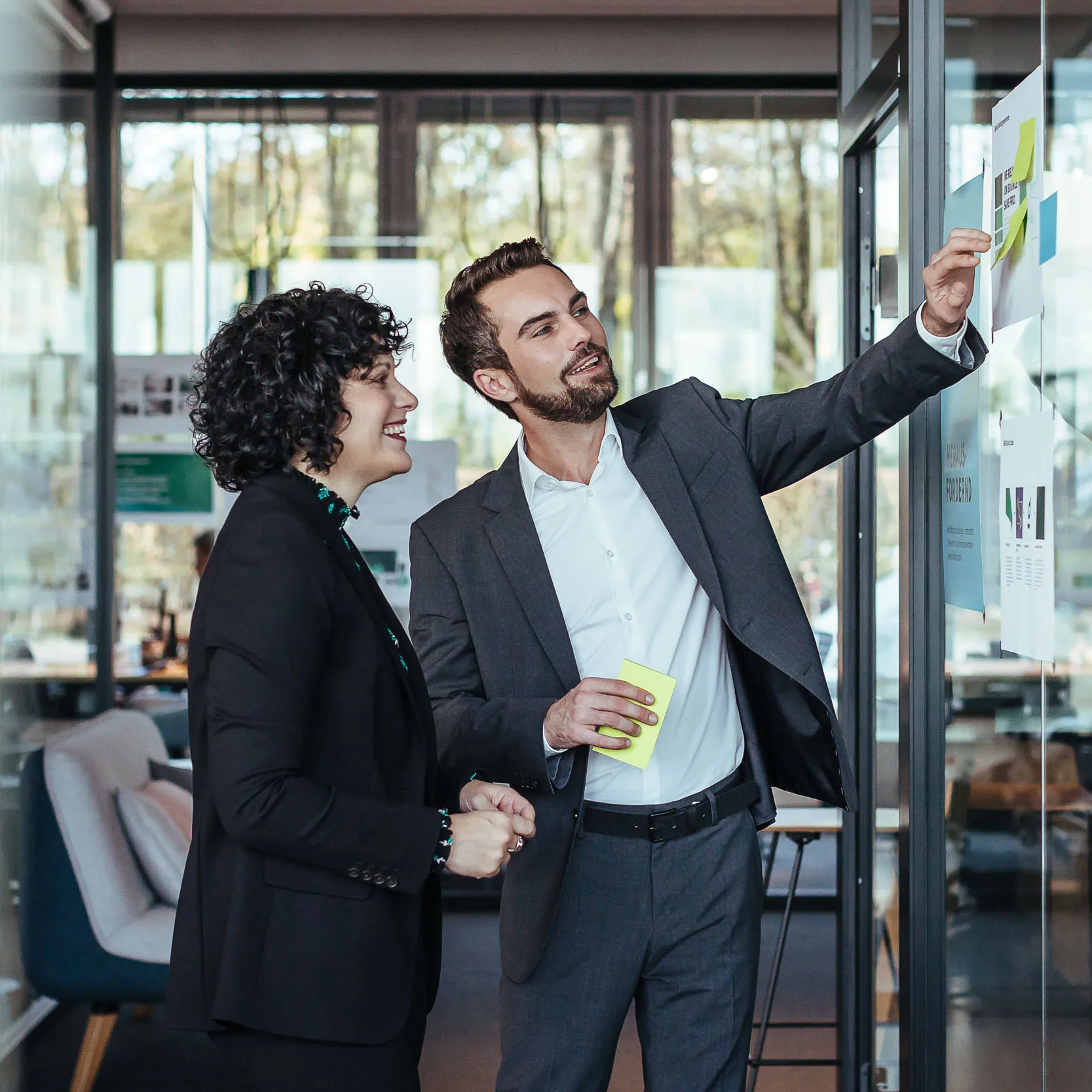 Zwei Geschäftspersonen, die in einem modernen Büro vor einer Glaswand mit Post-its und Dokumenten diskutieren.