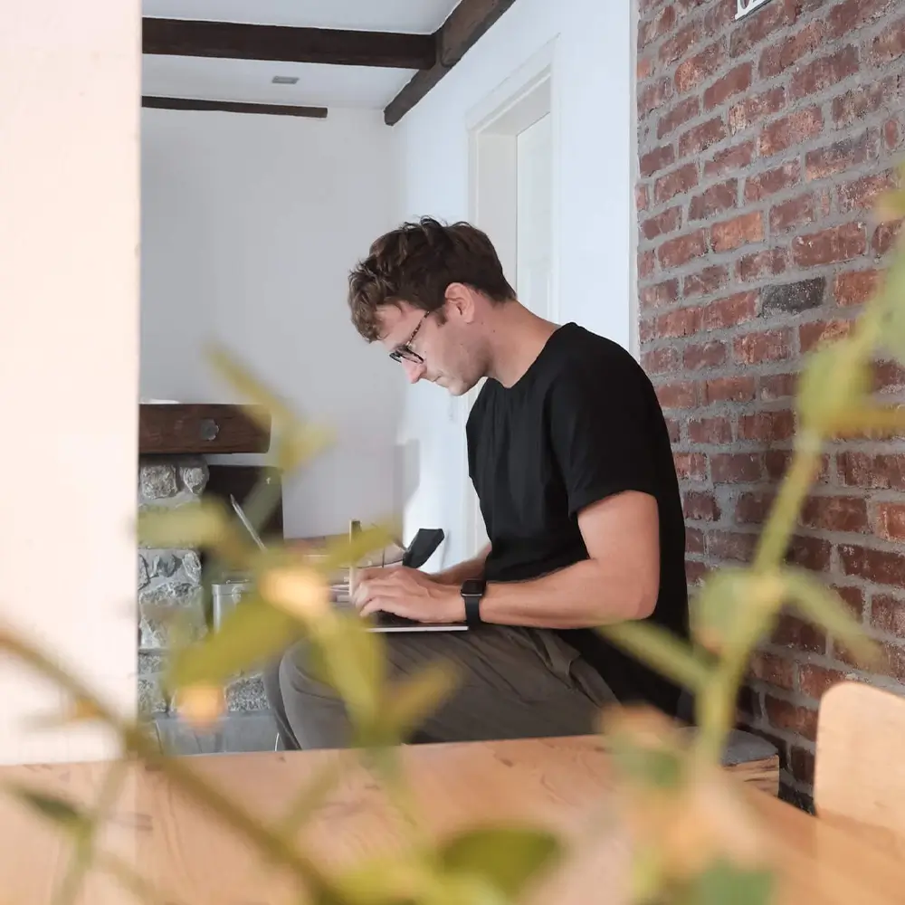 Hannes Eberlein, Gründer und Geschäftsführer von hej.build, arbeitet konzentriert an einem Tisch in einem Büro, umgeben von zwei Topfpflanzen. Das Szenario ist im Stil der urbanen Erkundung und minimalistischen Inszenierung gestaltet. Er entwickelt Konzepte, die Bauproduktherstellern bei der Zielgruppenansprache im Objektgeschäft helfen.