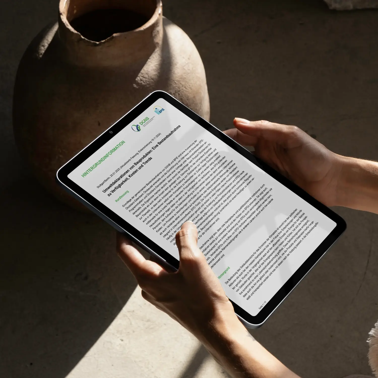 Hands hold a tablet displaying a digital document on Environmental Product Declarations (EPDs) against the backdrop of a ceramic vase. The tablet shows the heading »BACKGROUND INFORMATION« and a text on construction products and environmental data, while the natural ceramic contrasts with the digital content, symbolizing the connection between traditional building materials and modern sustainability data.