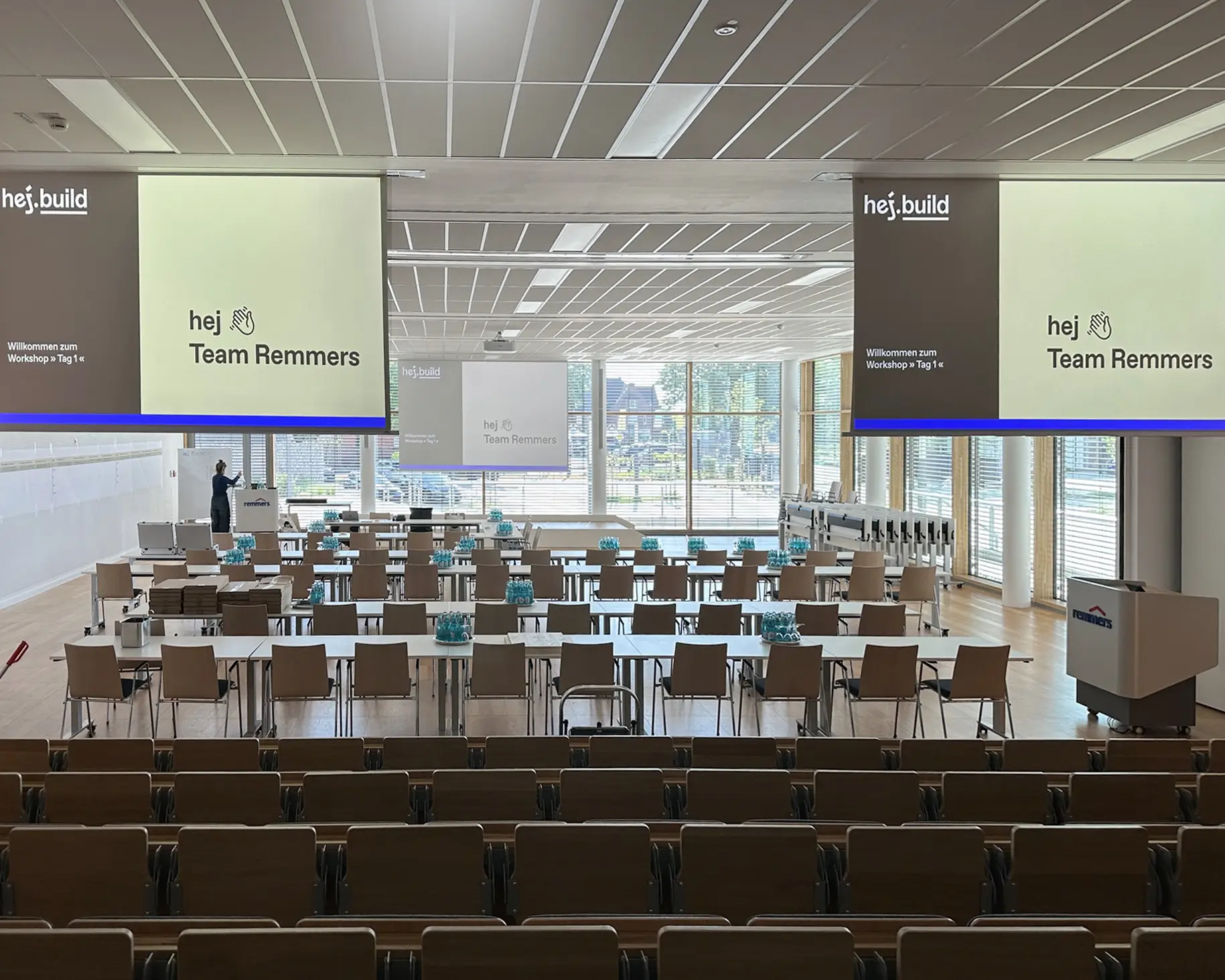 Blick in den vorbereiteten Workshop-Raum bei Remmers mit Präsentationsfolien „hej Team Remmers“ und Tischgruppen für den Auftakt-Workshop Tag 1.