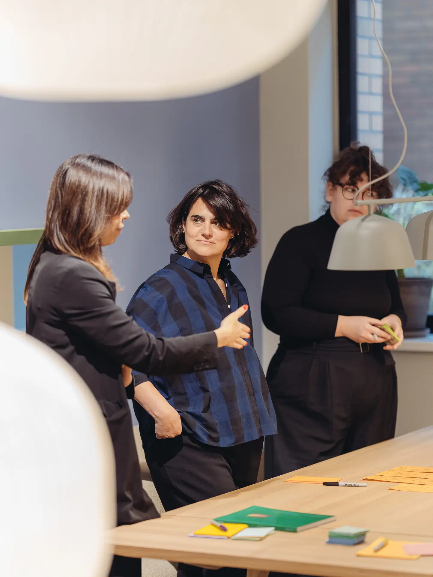 Three architects and designers in a creative workshop setting, discussing concepts at a table with note cards and documents, modern office with designer lighting in the background.