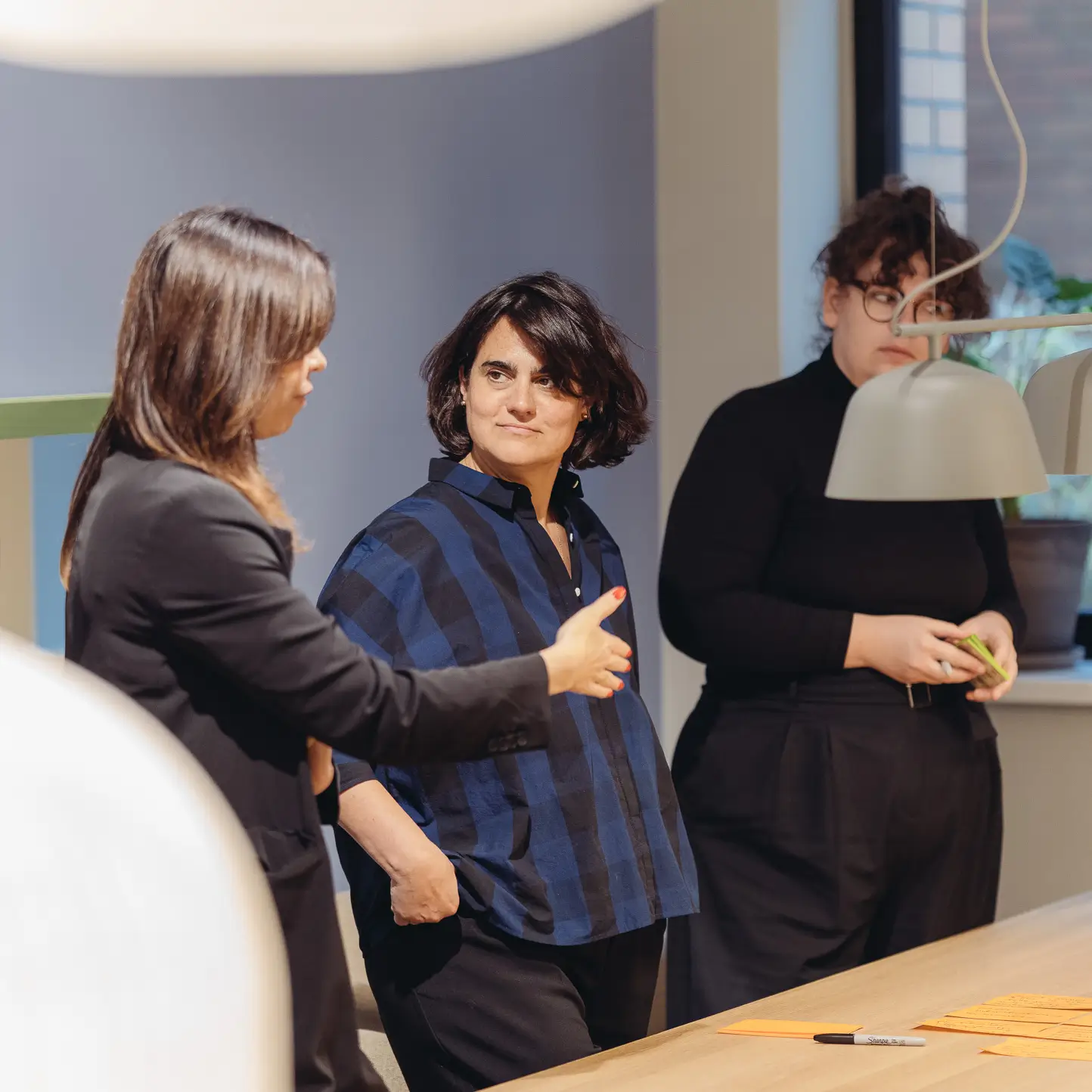 Three architects and designers in a creative workshop setting, discussing concepts at a table with note cards and documents, modern office with designer lighting in the background.