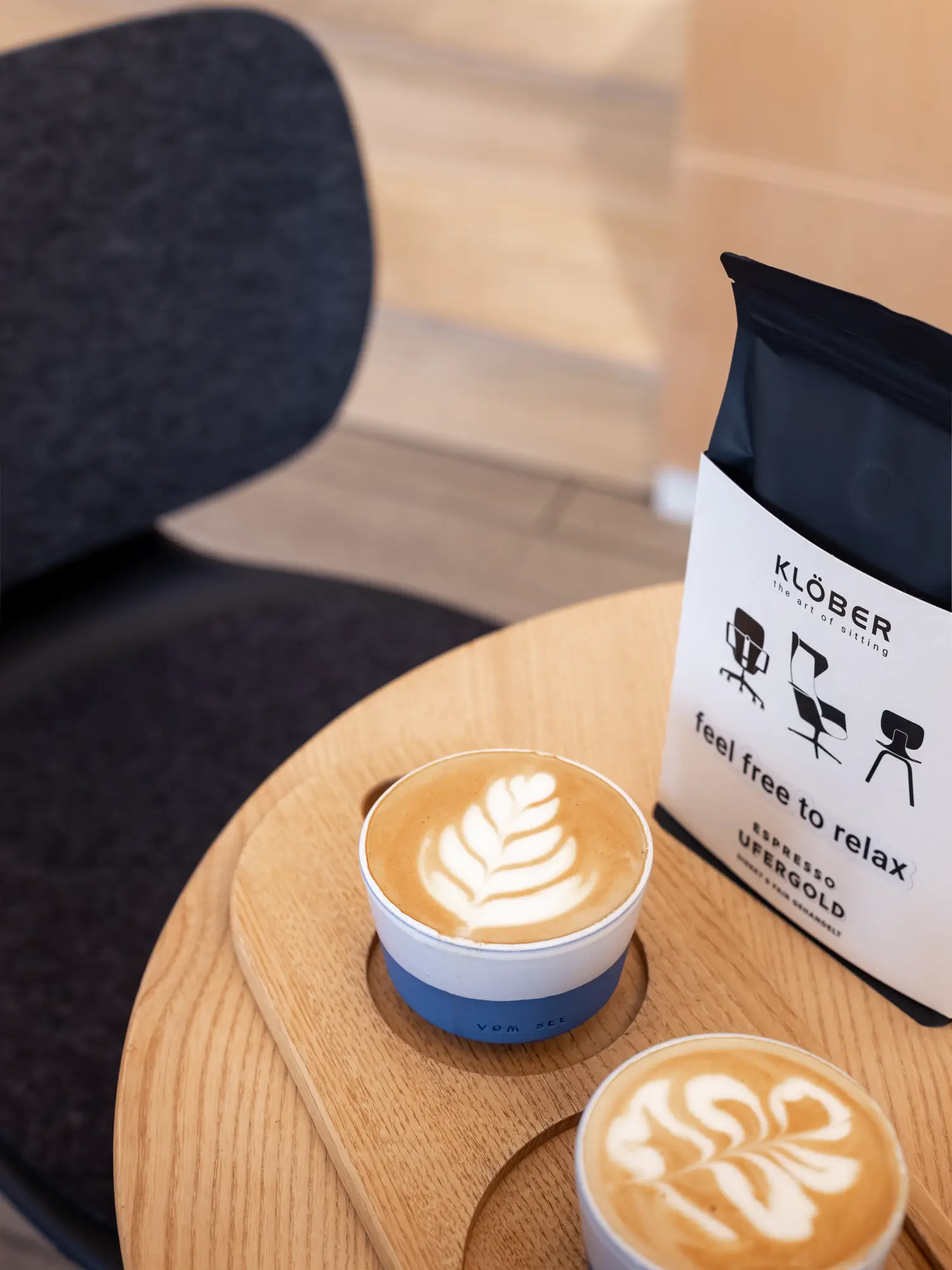 Two cappuccinos with latte art on a wooden table next to a package of Klöber espresso with the words "feel free to relax" – a modern coffee setup in a design environment.