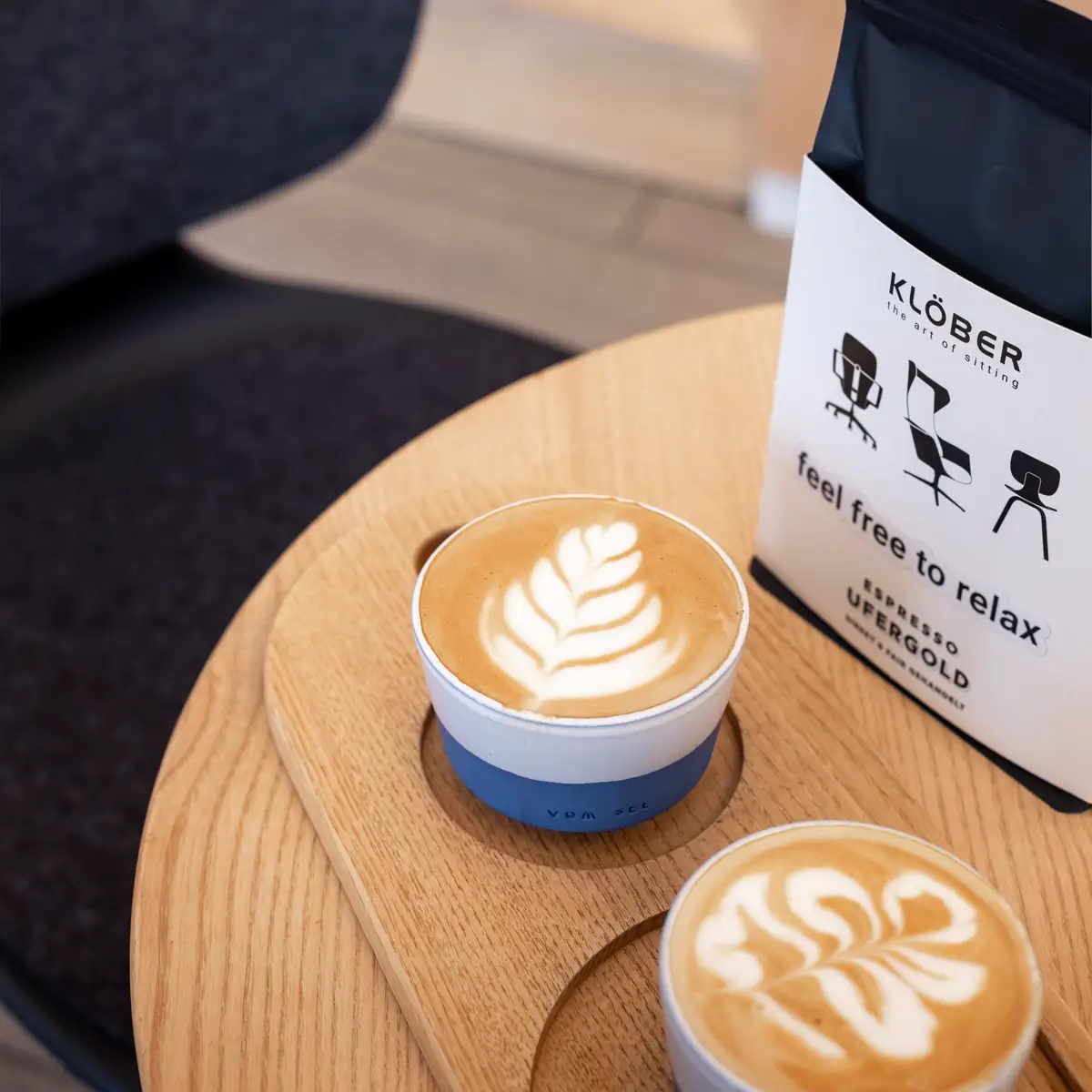Two cappuccinos with latte art on a wooden table next to a package of Klöber espresso with the words "feel free to relax" – a modern coffee setup in a design environment.