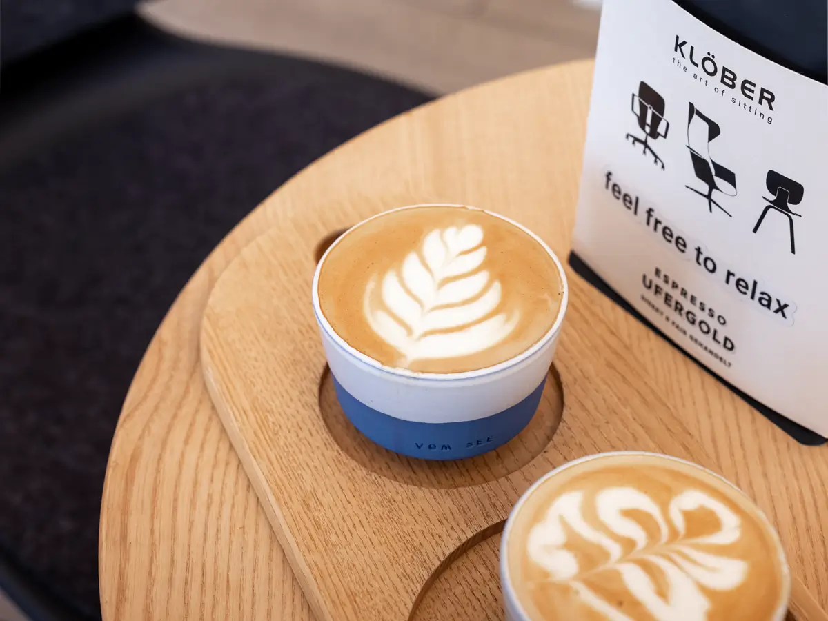 Zwei Cappuccinos mit Latte Art auf rundem Holztisch neben einer Packung Klöber Espresso ‚Ufergold‘, im modernen Lounge-Bereich mit Designerstuhl.
