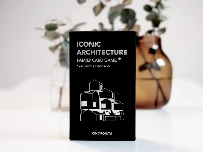Spielkartenset ‚Iconic Architecture Family Card Game‘ in schwarzer Verpackung mit grafischem Architekturmotiv, fotografiert vor minimalistischer Vase mit Eukalyptuszweigen.