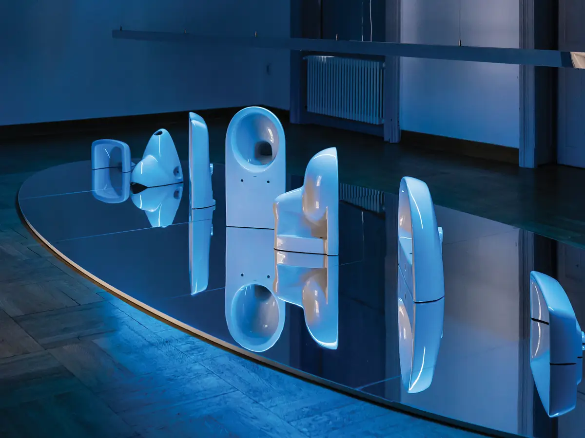 Installation „ROTA ET SPECULUM“ von Geberit, gestaltet vom hanczar studio: Sanitärkeramik wie Toiletten und Urinale sind auf einer großen Spiegelplatte arrangiert und in blauem Licht inszeniert.