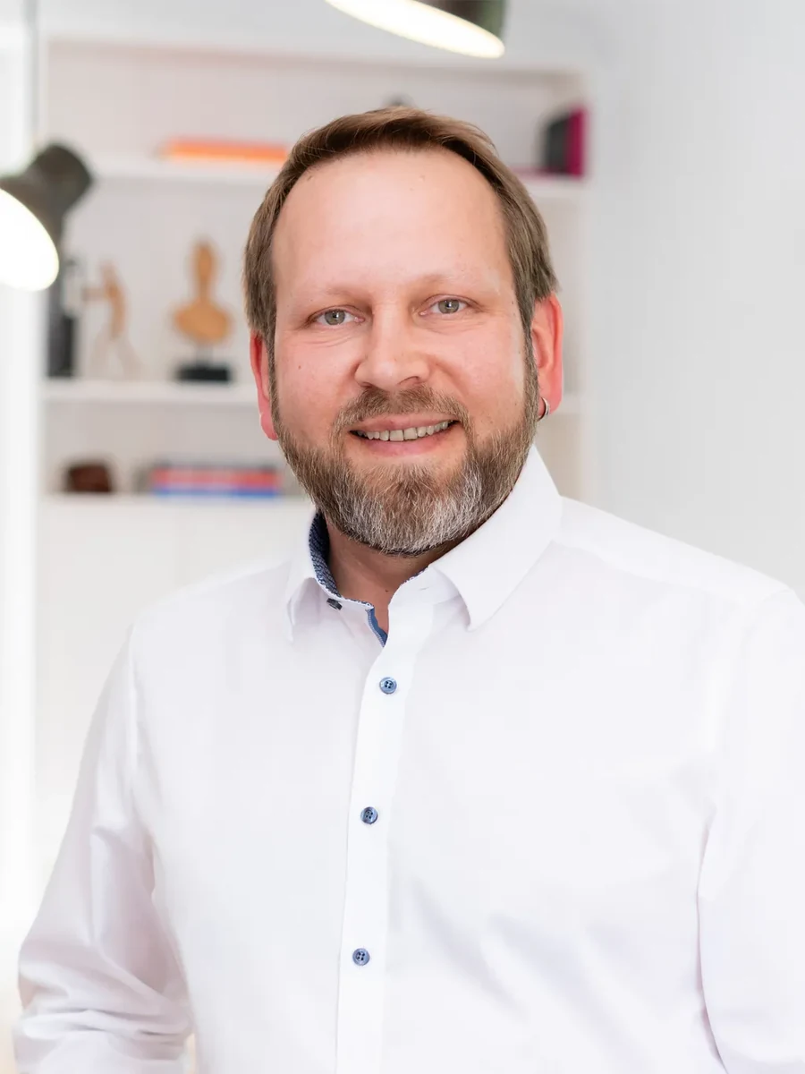 Heinrich Böhm von JOI-Design Innenarchitekten, lächelnd in einem weißen Hemd vor einem hellen Bürohintergrund.