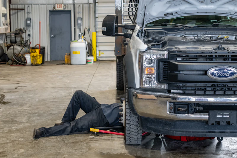 A mechanic lies on the floor beneath the front of a white Ford pickup truck with its hood open inside a vehicle repair shop, tools scattered nearby as they work under the engine.