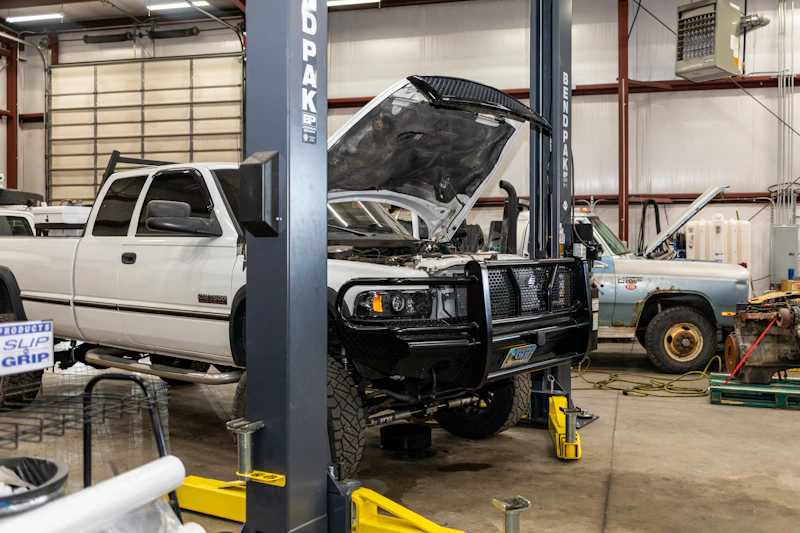 Wheel alignment service with pickup truck lifted on hydraulic hoist, hood open for inspection in heavy-duty repair shop, tools and another truck visible in background.