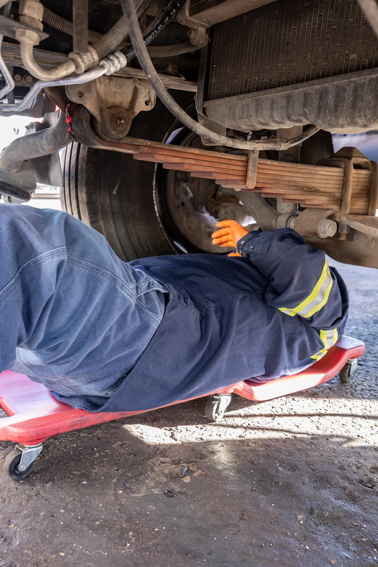 Technician performs preventive maintenance beneath a heavy-duty truck, inspecting the suspension and wheel assembly.