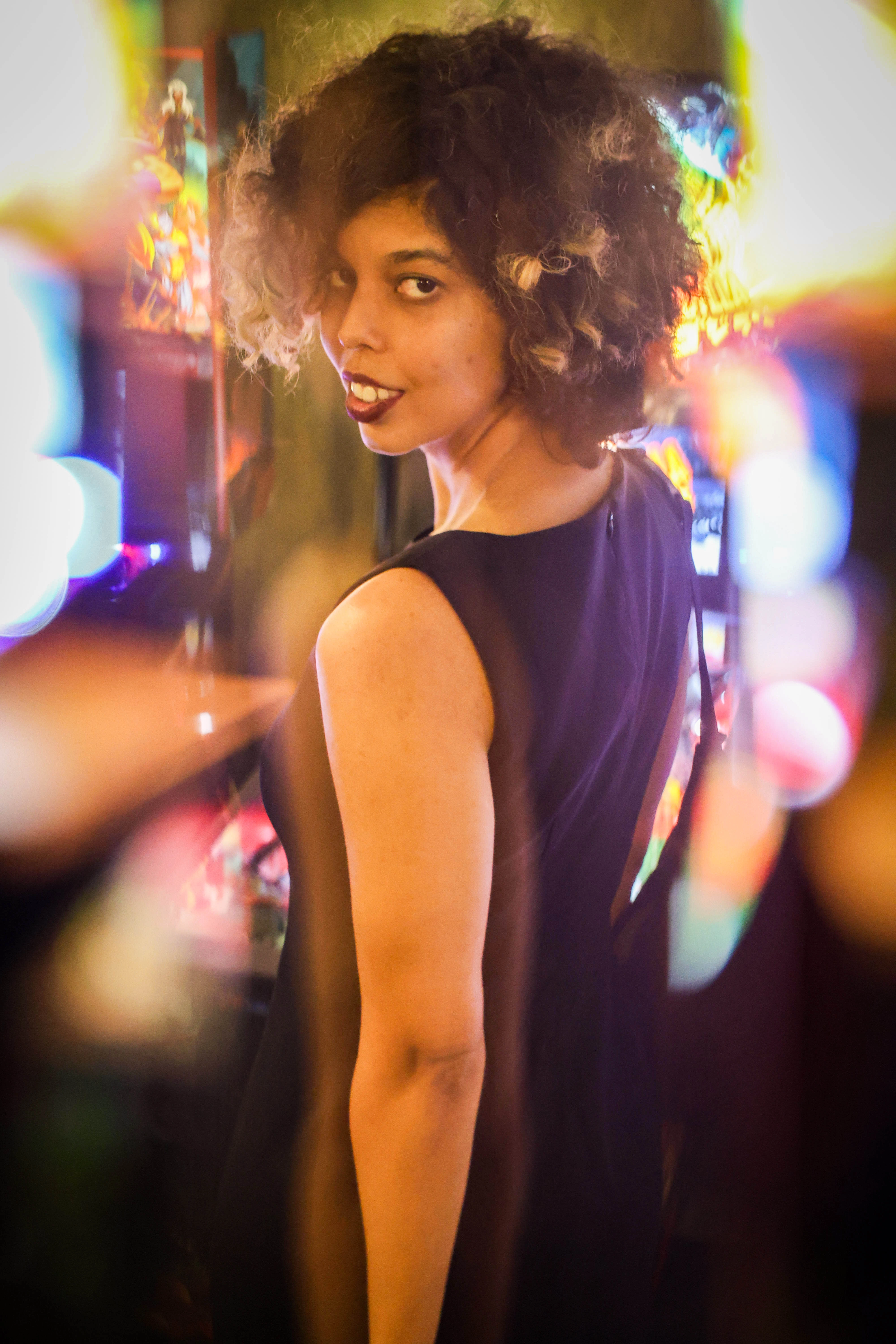 A photo of Amelia Som, a tan skinned person in a black dress. They have curly neck-length black hair with blonde tips. They have a slight smile to their face and are backlit by colorful arcade lights.