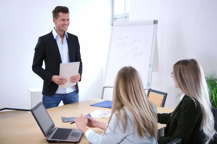Formateur souriant présente un cours à deux participantes attentives, avec un tableau blanc en arrière-plan, dans une session de formation en investissement immobilier.