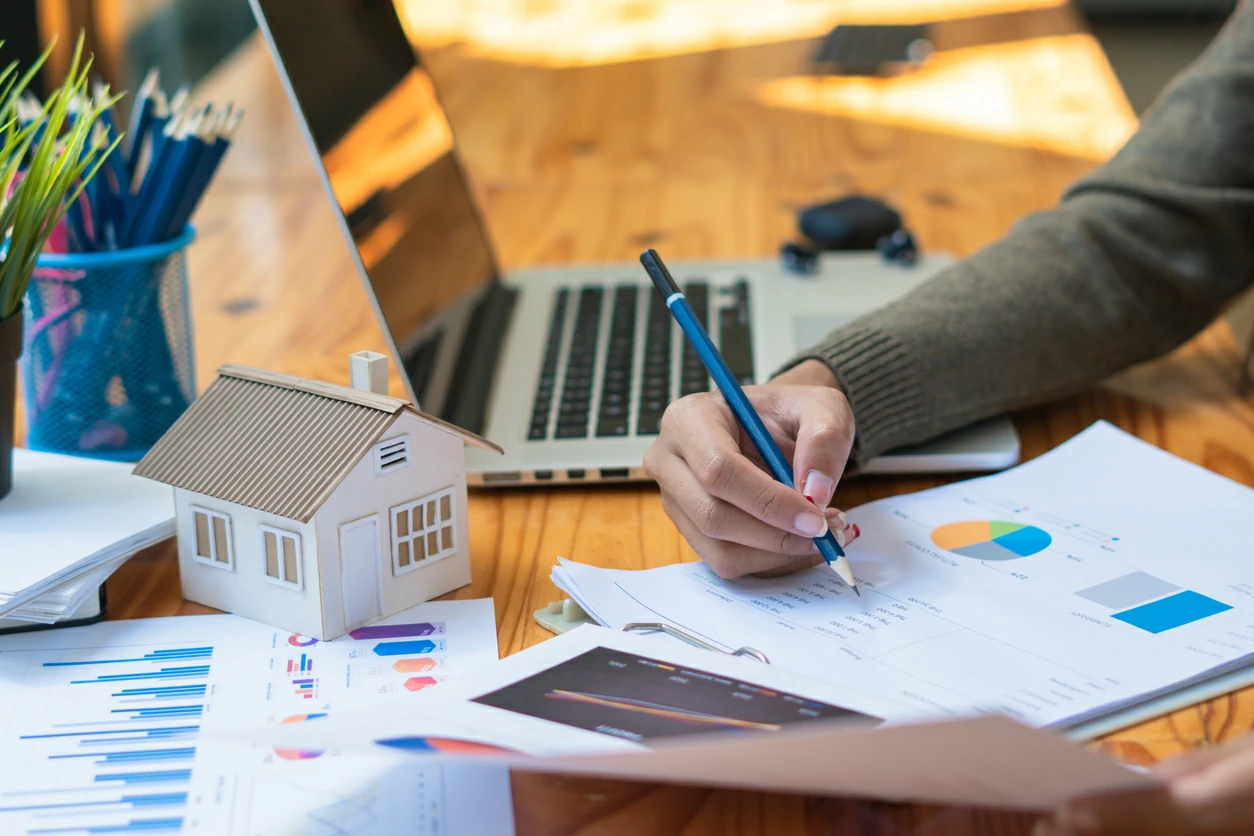 Homme consultant des documents chiffrés avec un crayon à côté d'une maquette de maison et d'un ordinateur portable.