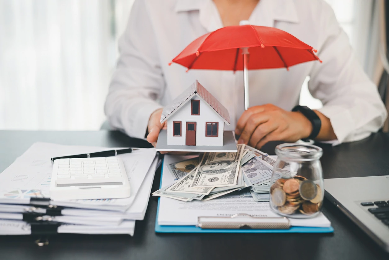 Homme tenant une petite maison et un petit parapluie devant des pièces de monnaie et des liasses de billet.