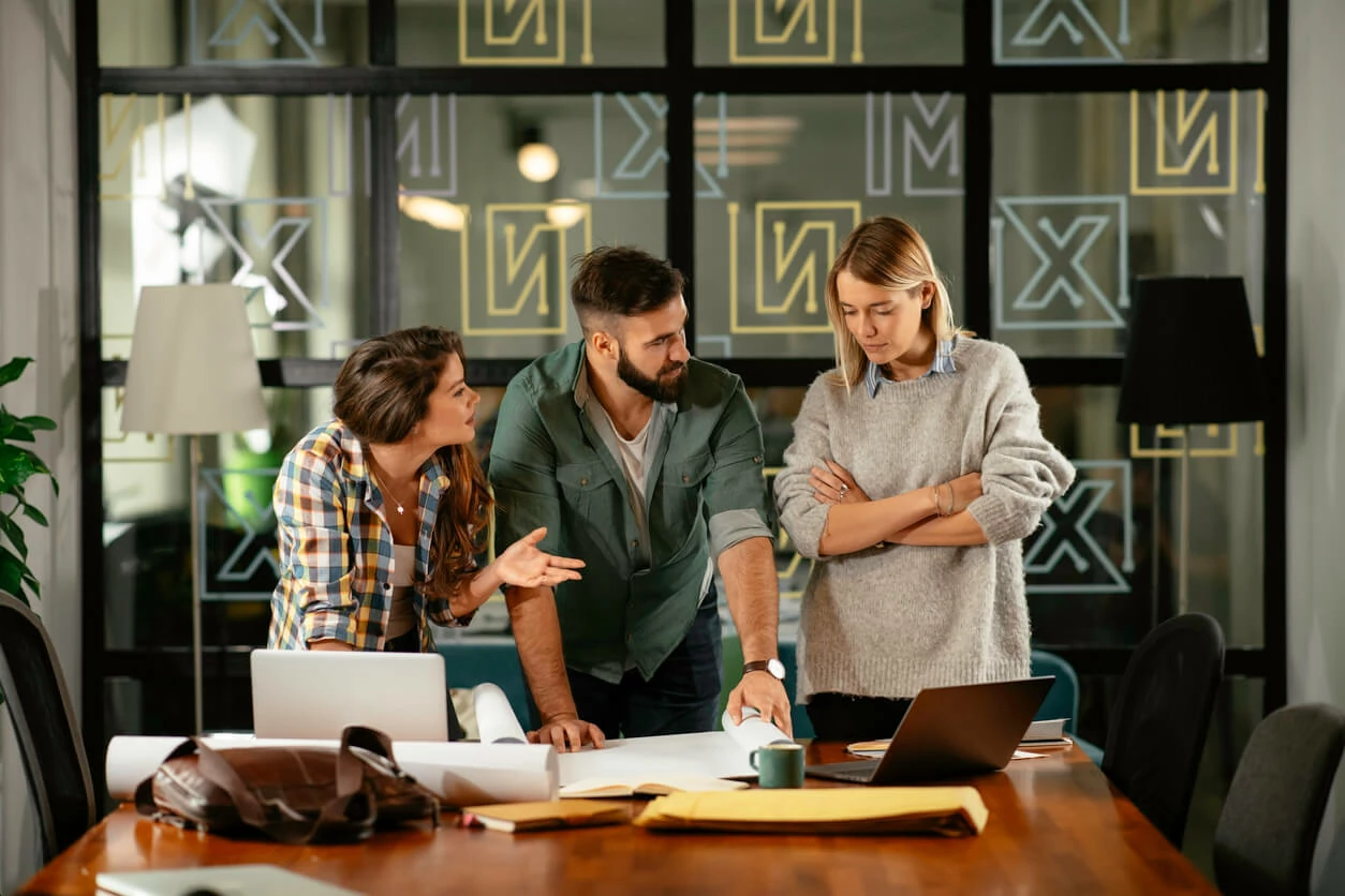 Un homme et deux femmes en pleine concertation devant des ordinateurs portables et des documents dans une salle de réunion.e