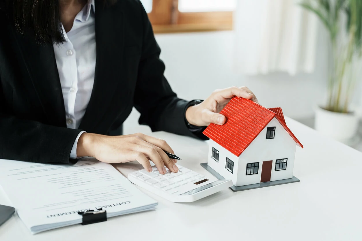 Femme utilisant une calculatrice d'une main et tenant une petite maison de l'autre main à côté d'un document.