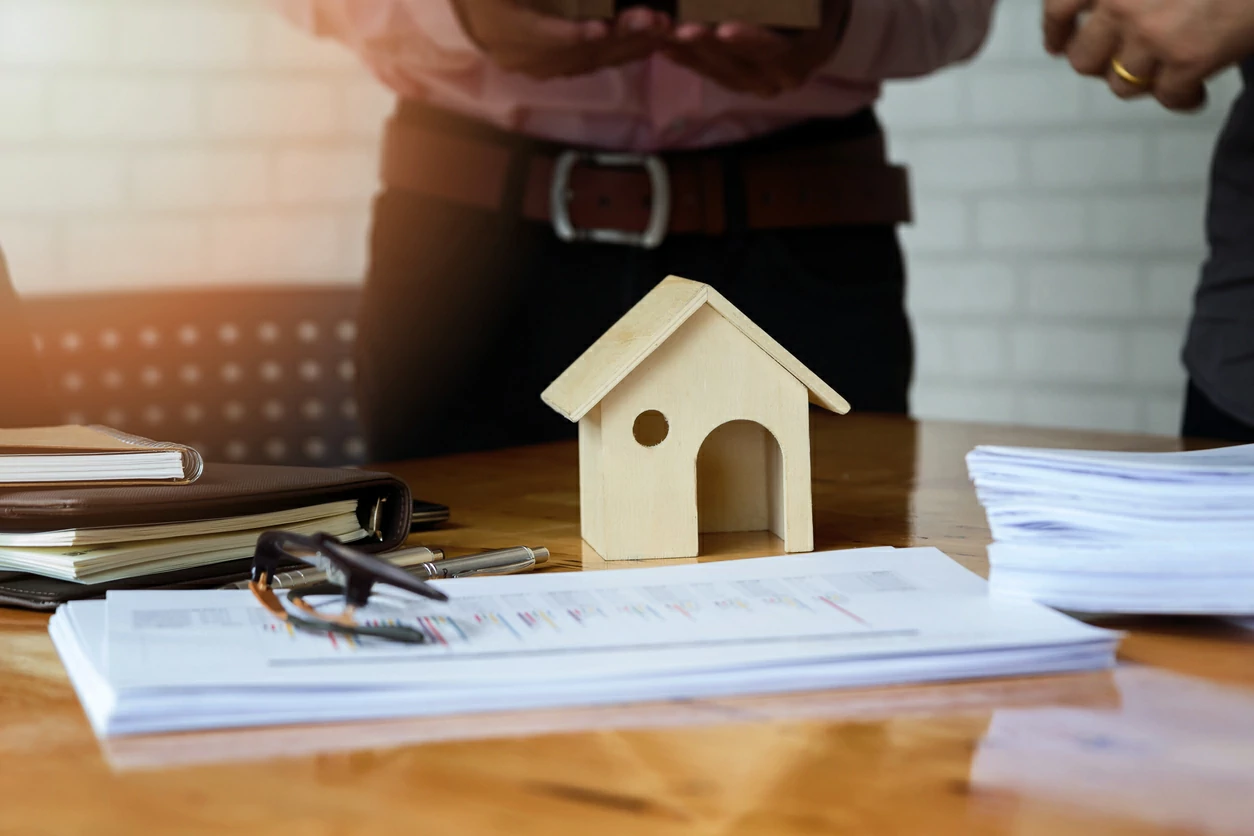 Petite maison en bois posée au-dessus d'une pile de documents et deux hommes discutant en arrière-plan..