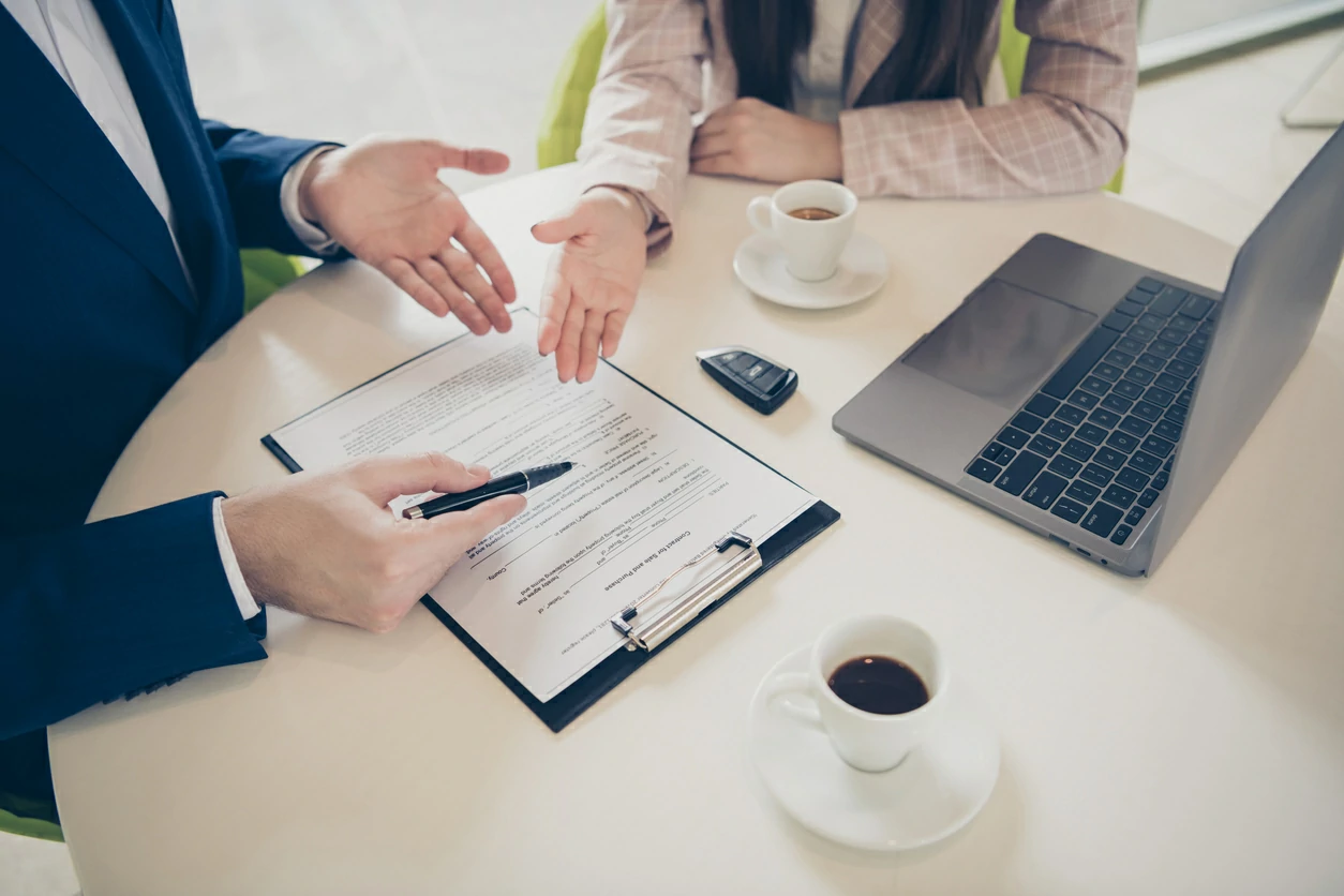 Un homme et une femme consultant un document à coté d'un ordinateur portable, deux tasses de café et une clé de voiture.