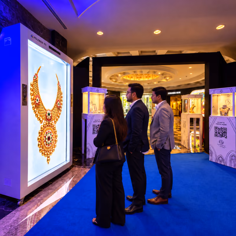 Visitors at a HoloBox911 display showing a gold necklace set beside exhibitor cabinets at COUTURE INDIA 2026, New Delhi