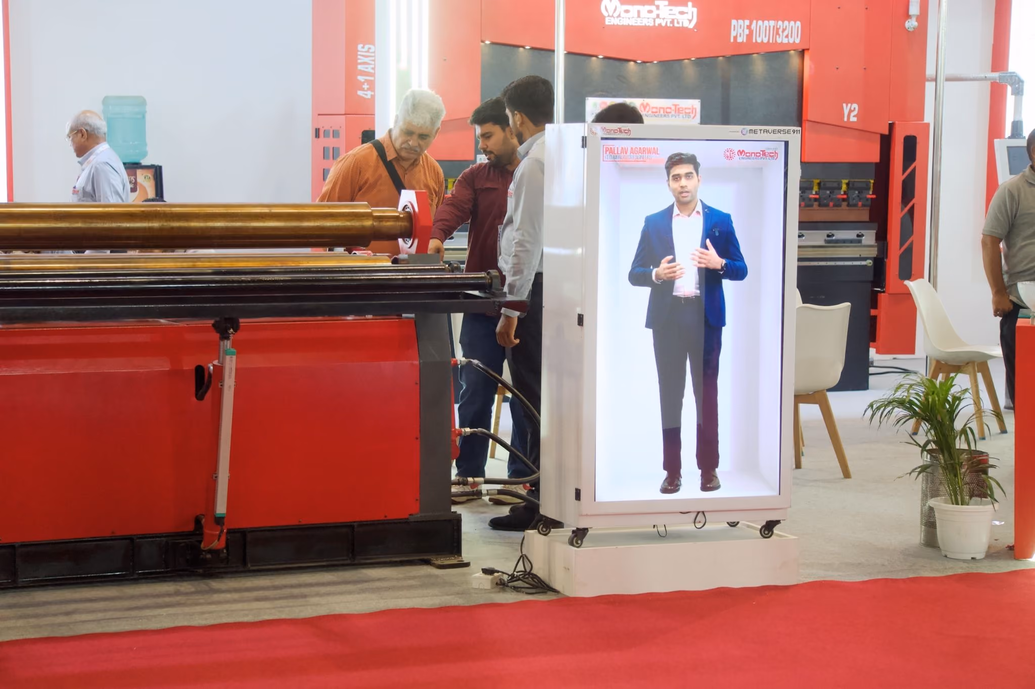 Visitors at the Monotech Engineers stand at IMTOS 2025, with the HoloBox911 unit playing the recorded welcome beside red sheet metal machinery on the show floor.