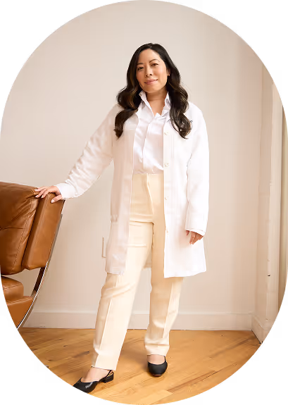 Woman with long dark hair wearing a white coat, white shirt, cream pants, and black shoes standing indoors next to a brown leather chair.