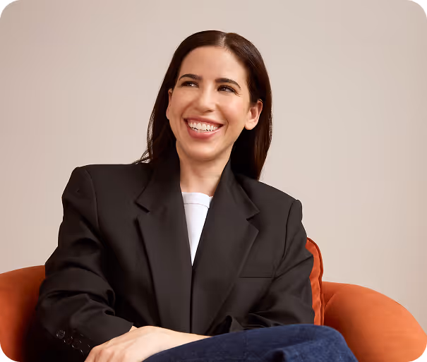 Smiling woman with long dark hair wearing a black blazer and white shirt sitting on an orange chair against a beige background.