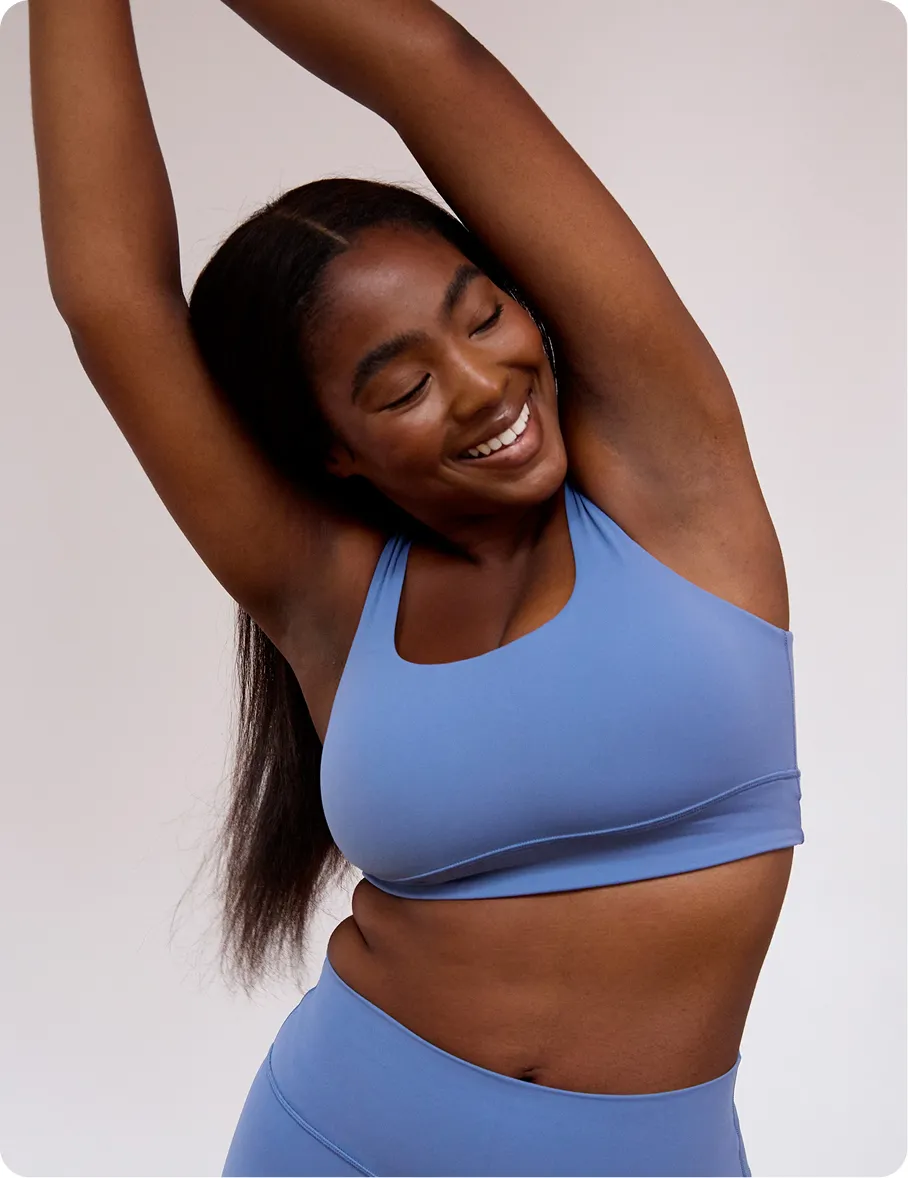 Smiling woman in blue athletic wear stretching with arms raised against a neutral background.