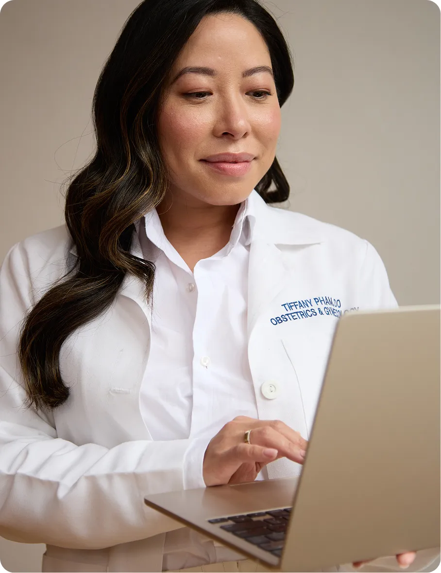 Female doctor in white coat using a laptop, with name and specialty embroidered on the coat.