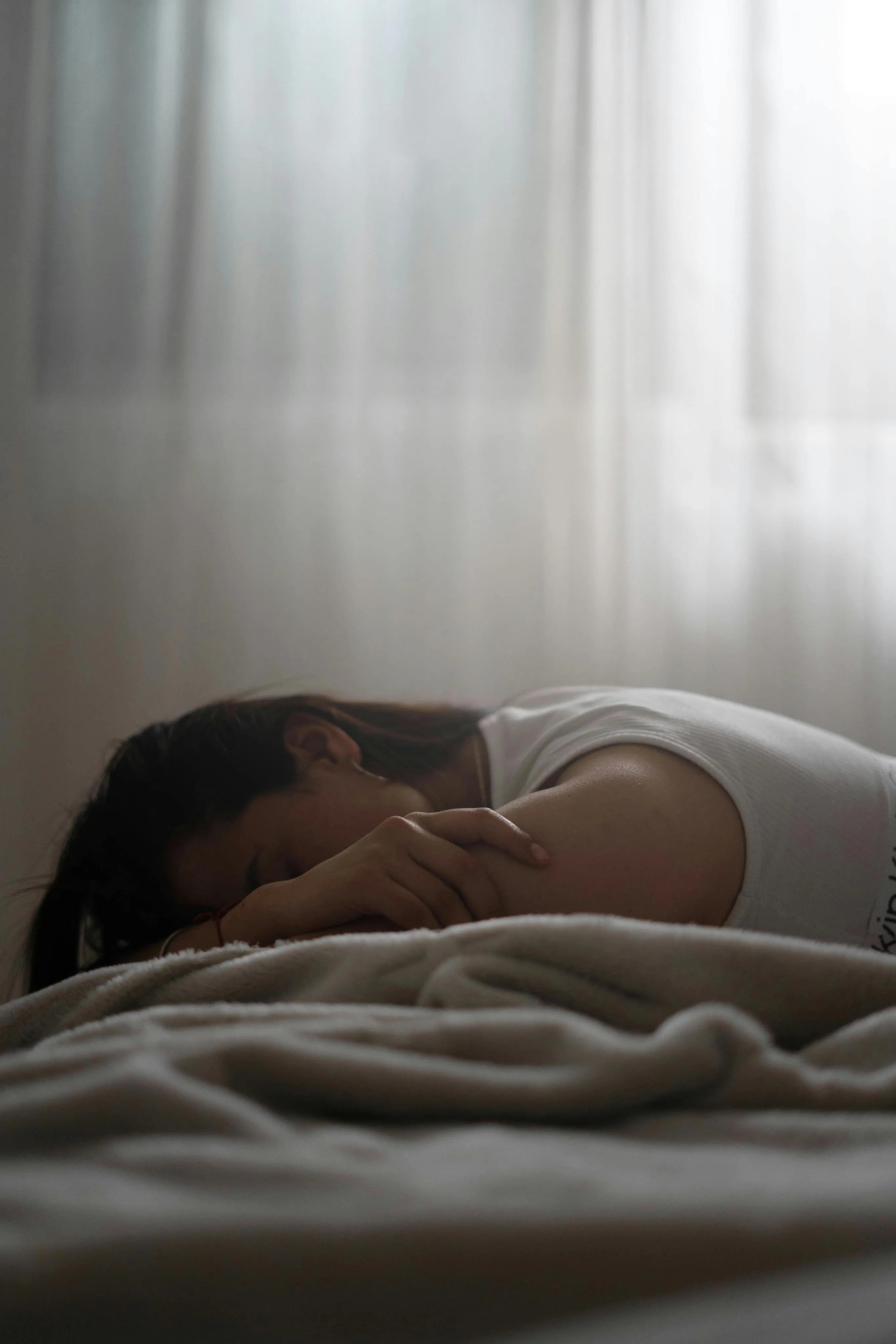 Person lying face down on a bed with soft lighting and sheer curtains in the background.