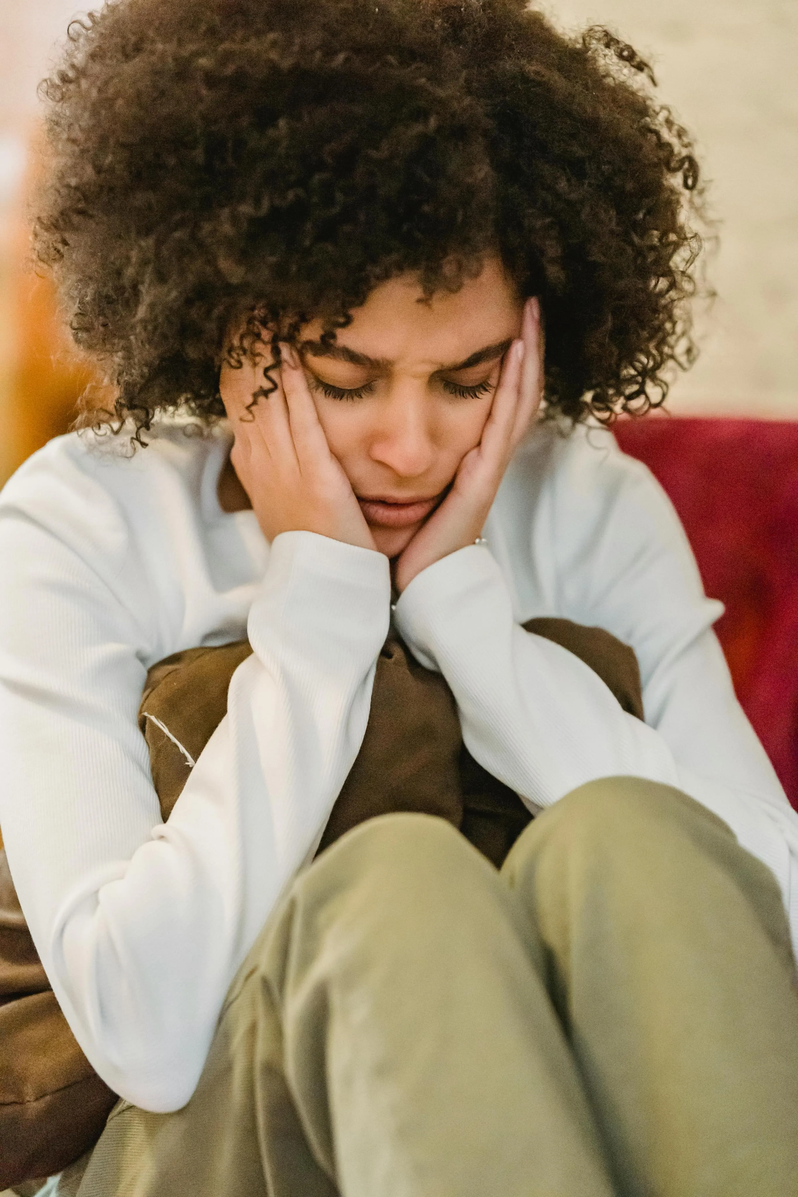 Young woman with curly hair sitting with her knees up, holding her face and looking distressed.