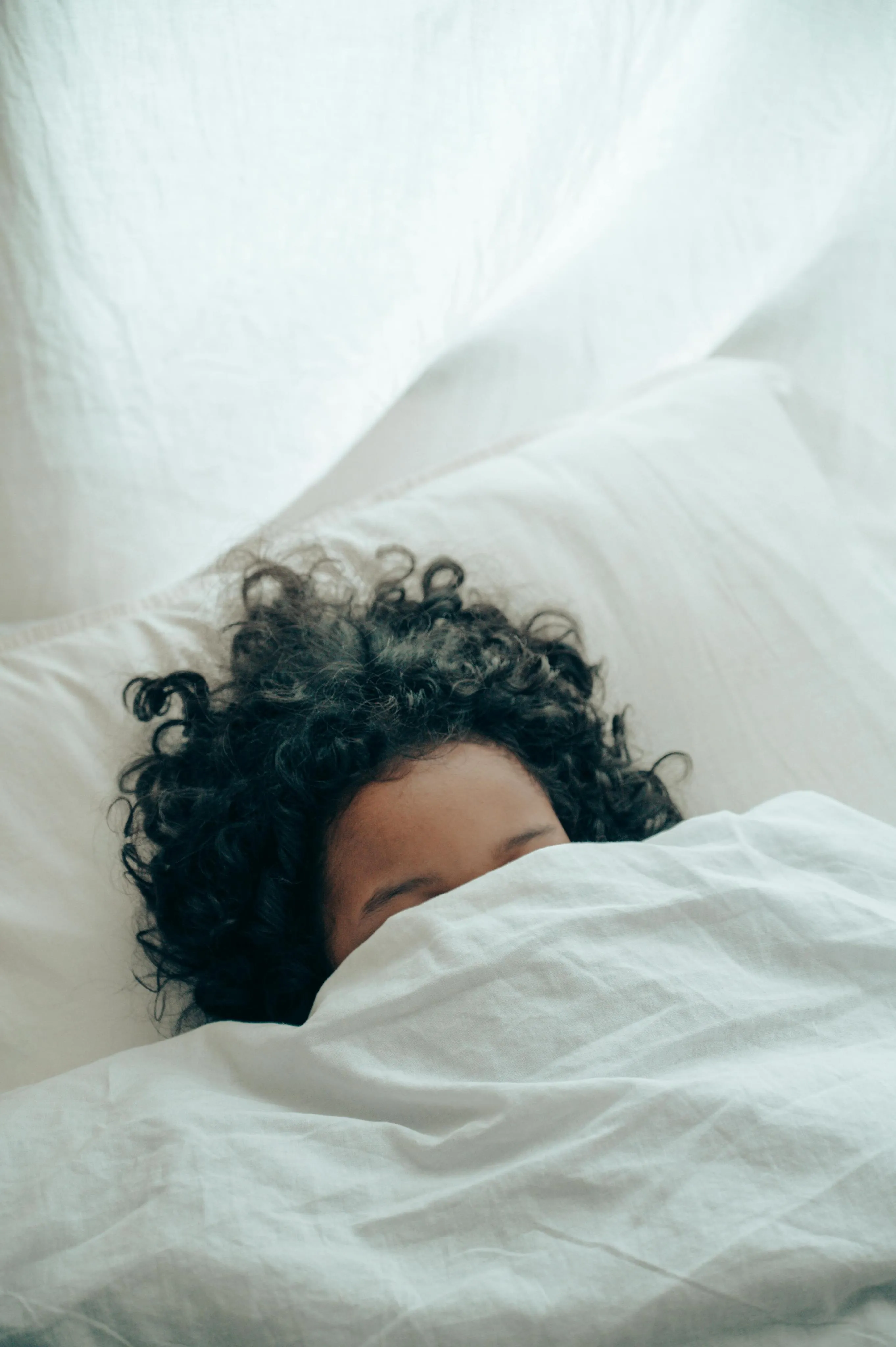 Person with curly hair partially covered by white blanket lying on white bed sheets.