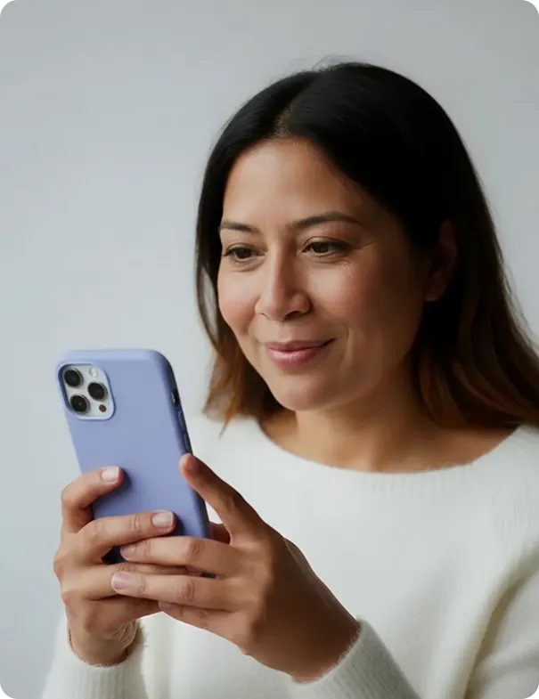 Woman in a white sweater looking and smiling at a smartphone with a purple case.