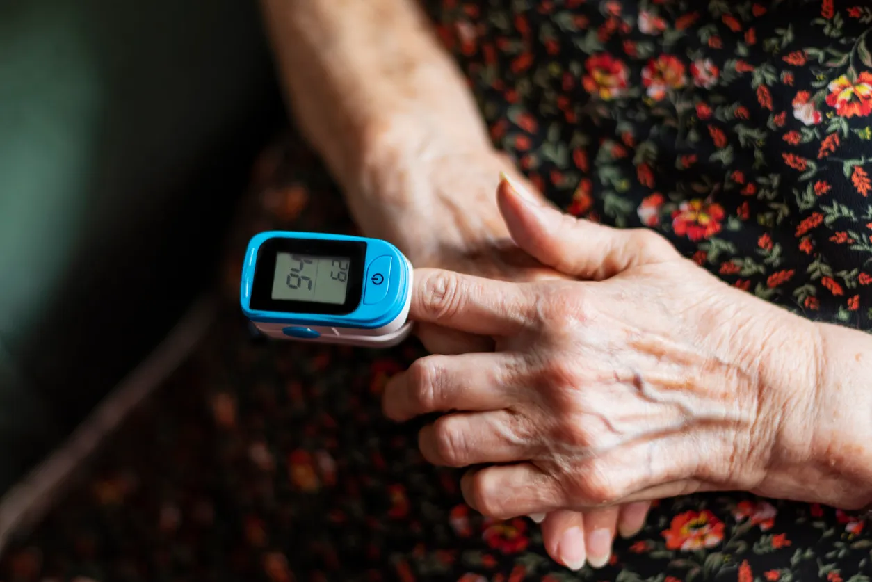 Elderly person’s hand with a blue fingertip pulse oximeter displaying oxygen saturation (97) and pulse rate (69).