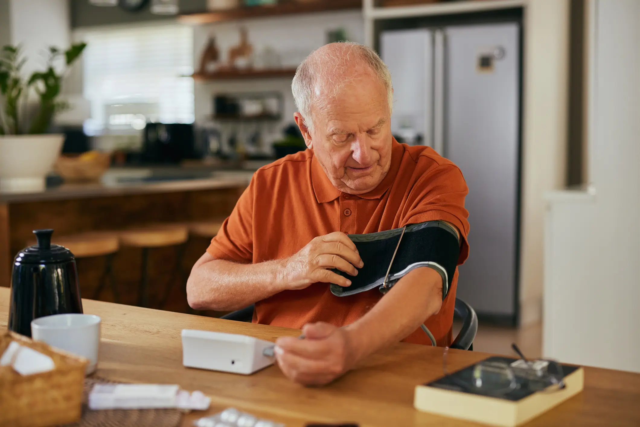 Older man in orange polo shirt using a blood pressure cuff and monitor at home