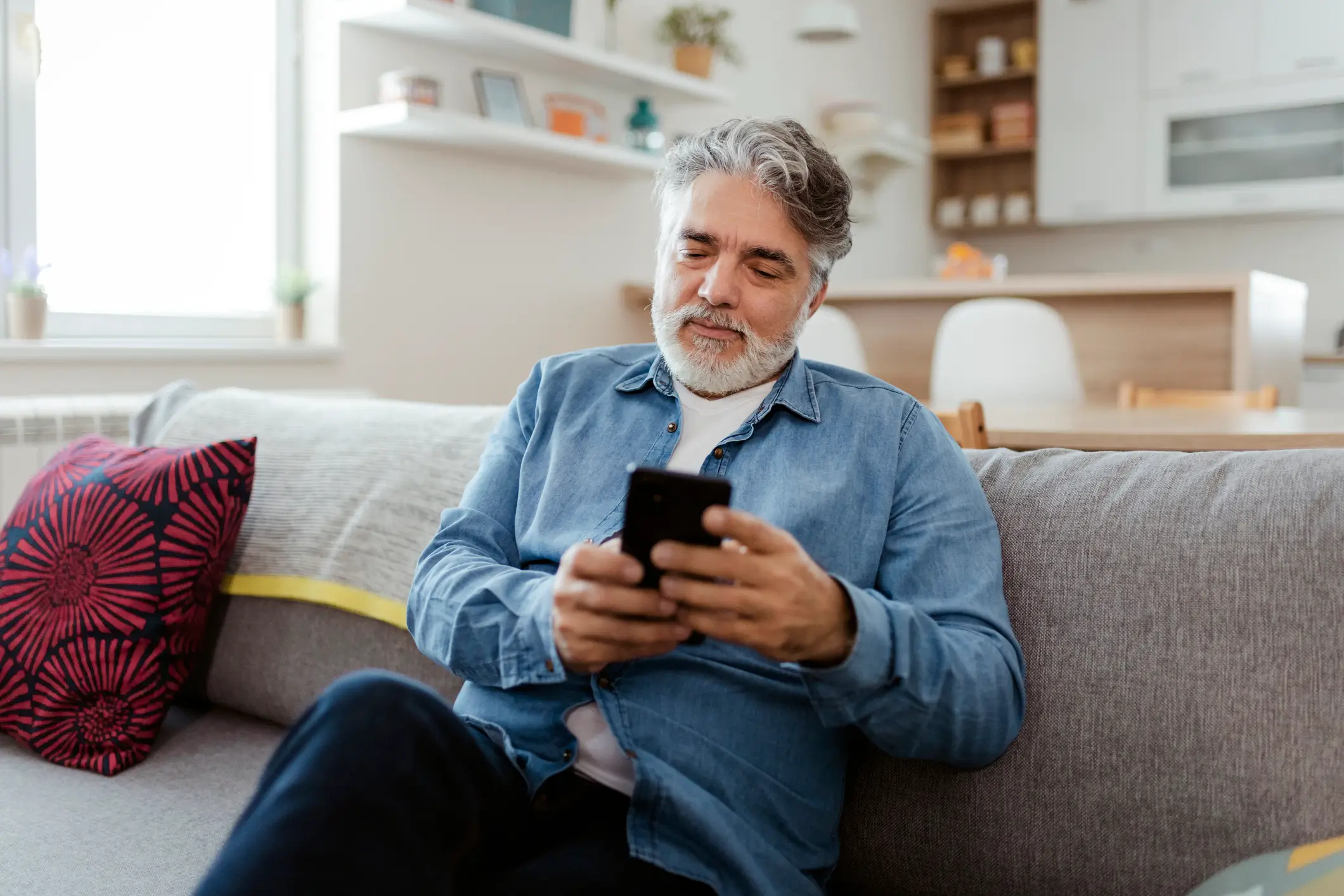 Portrait of bearded mature man wearing casual clothes sitting on a couch at the living room, using mobile phone