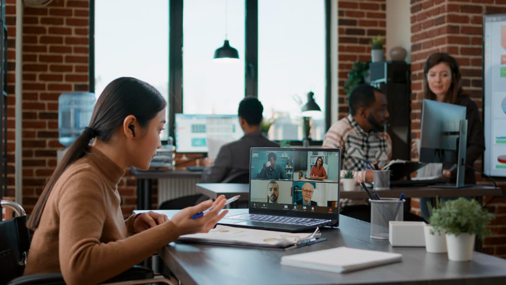 Une femme est assise au bureau avec son équipe hybride et participe à une réunion virtuelle.