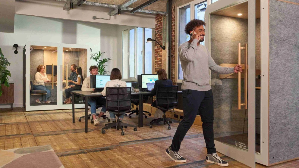 Ein Mann telefoniert in einem vollen Büro und geht währenddessen in eine Telefonbox.