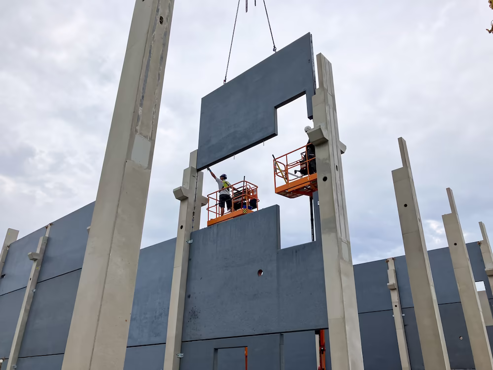 Two construction workers in lift platforms installing a large precast concrete wall panel between tall structural columns.