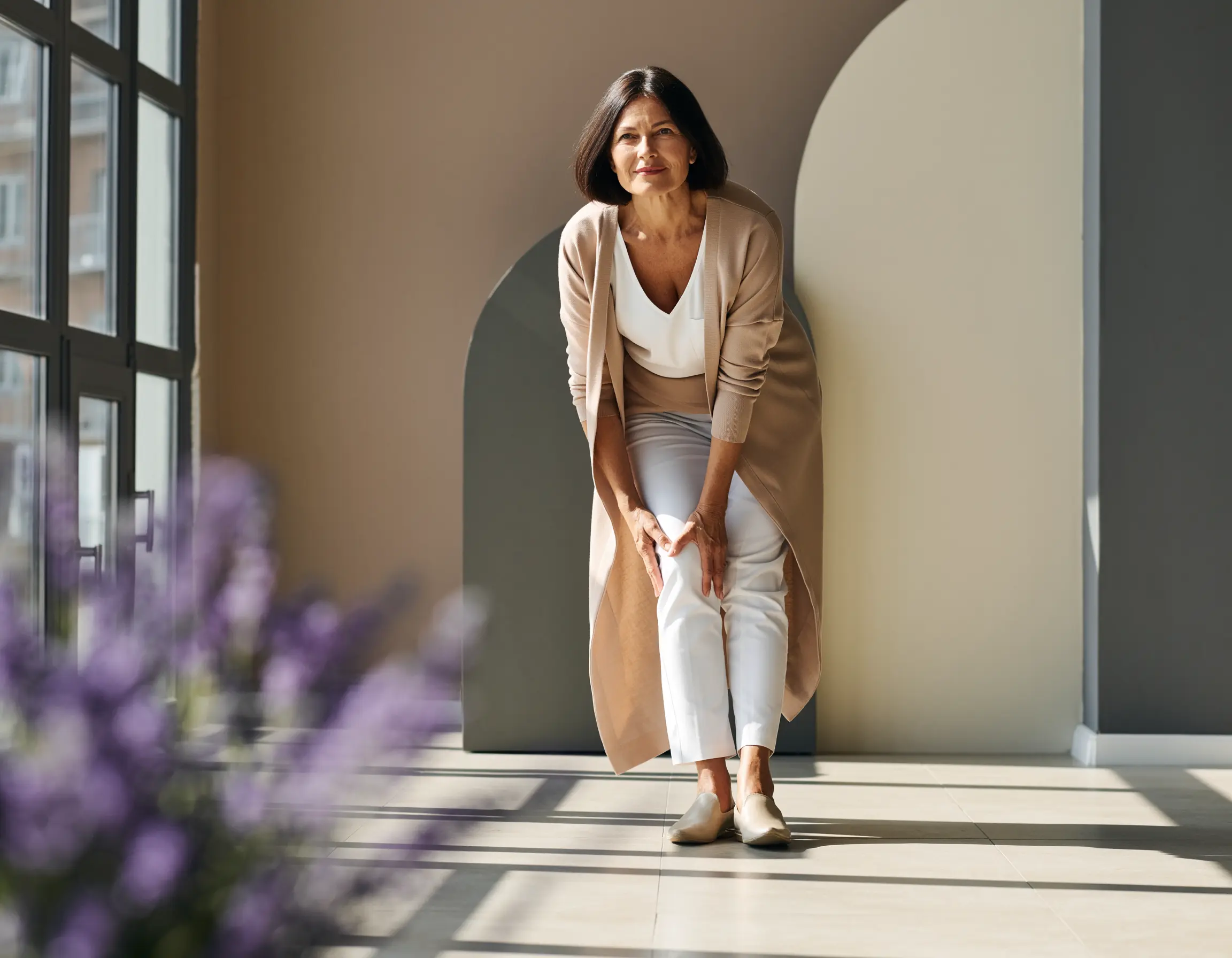 Middle-aged woman bent forward holding her right knee, standing in a sunlit room with large windows and blurred purple flowers in the foreground.
