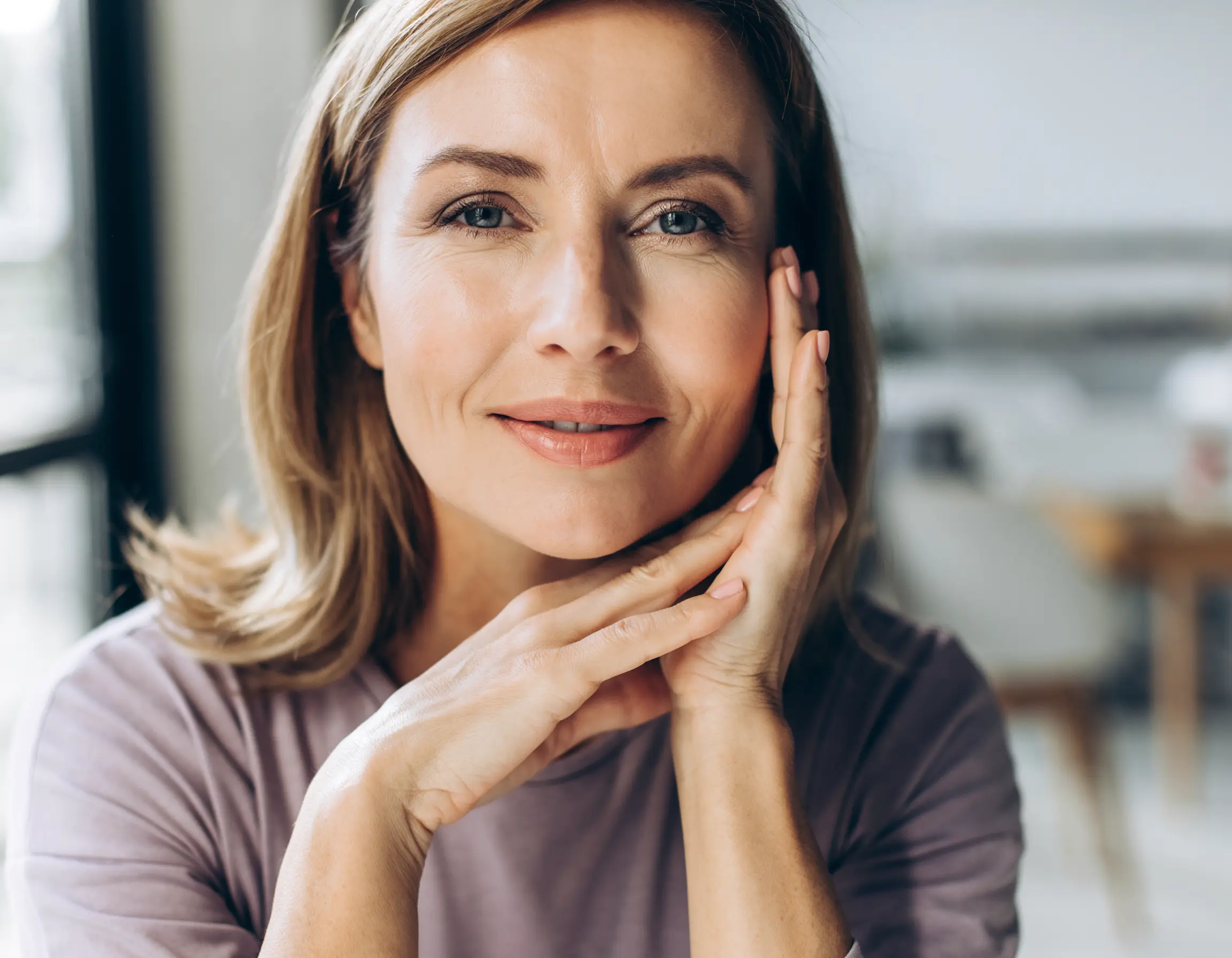 Close-up portrait of a smiling middle-aged woman with light makeup, resting her face on her hands indoors.