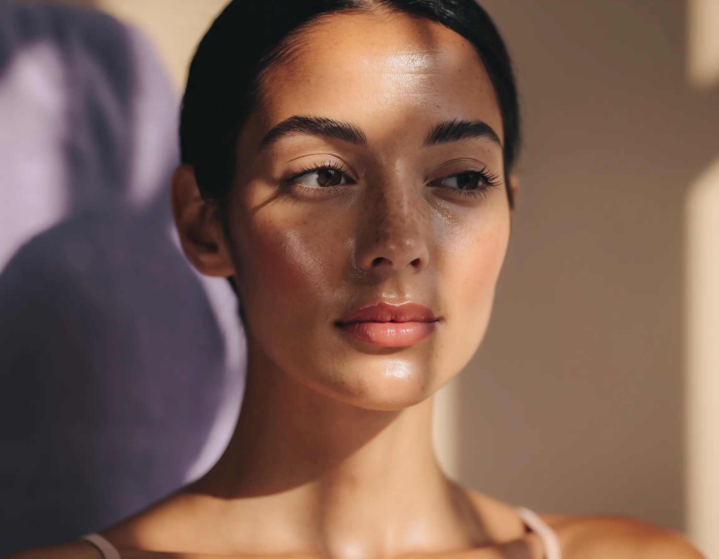 Close-up of a woman with smooth skin, subtle freckles, and natural makeup, looking to the side with soft shadows on her face.
