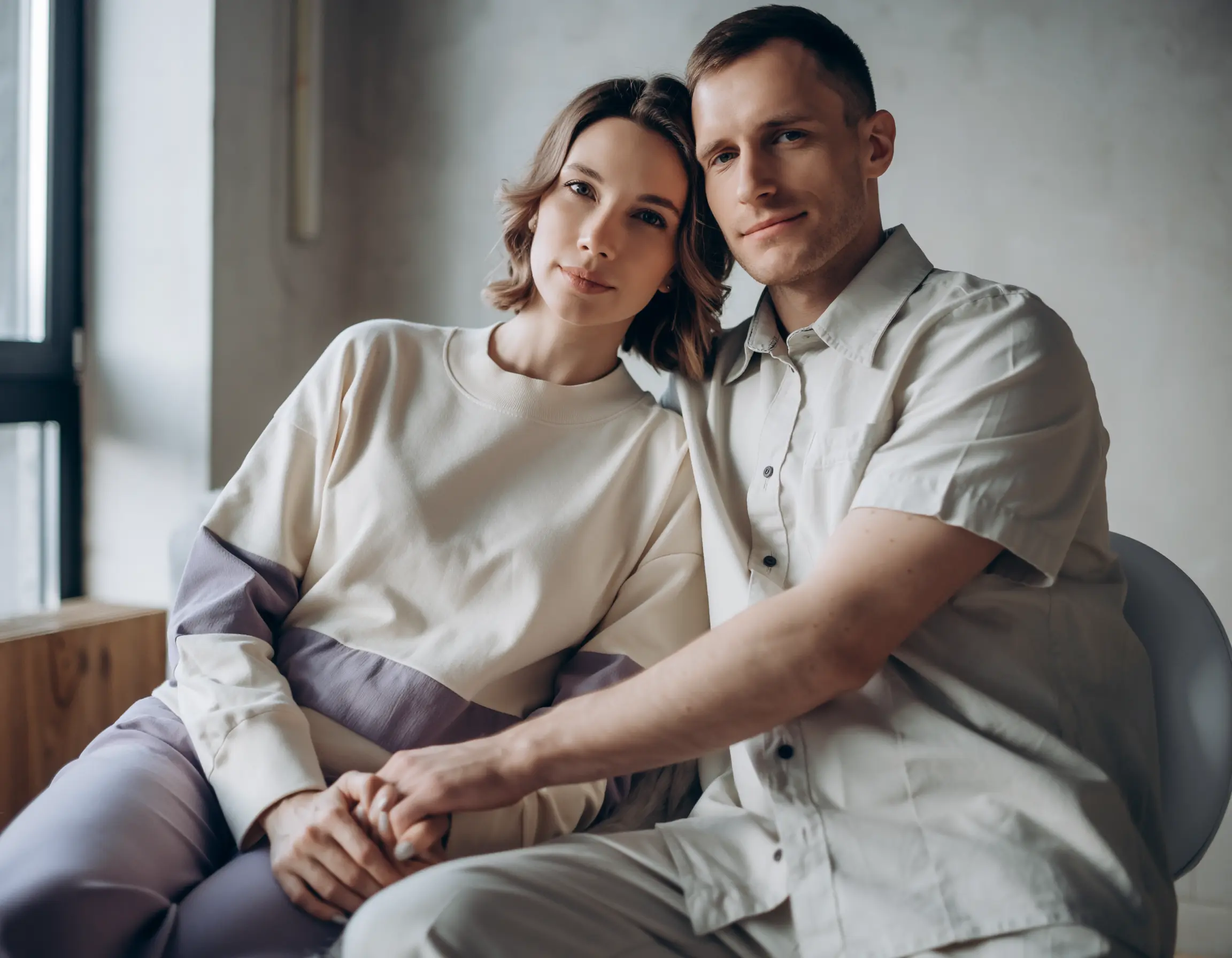 Young couple sitting closely indoors, holding hands and looking at the camera with gentle expressions.